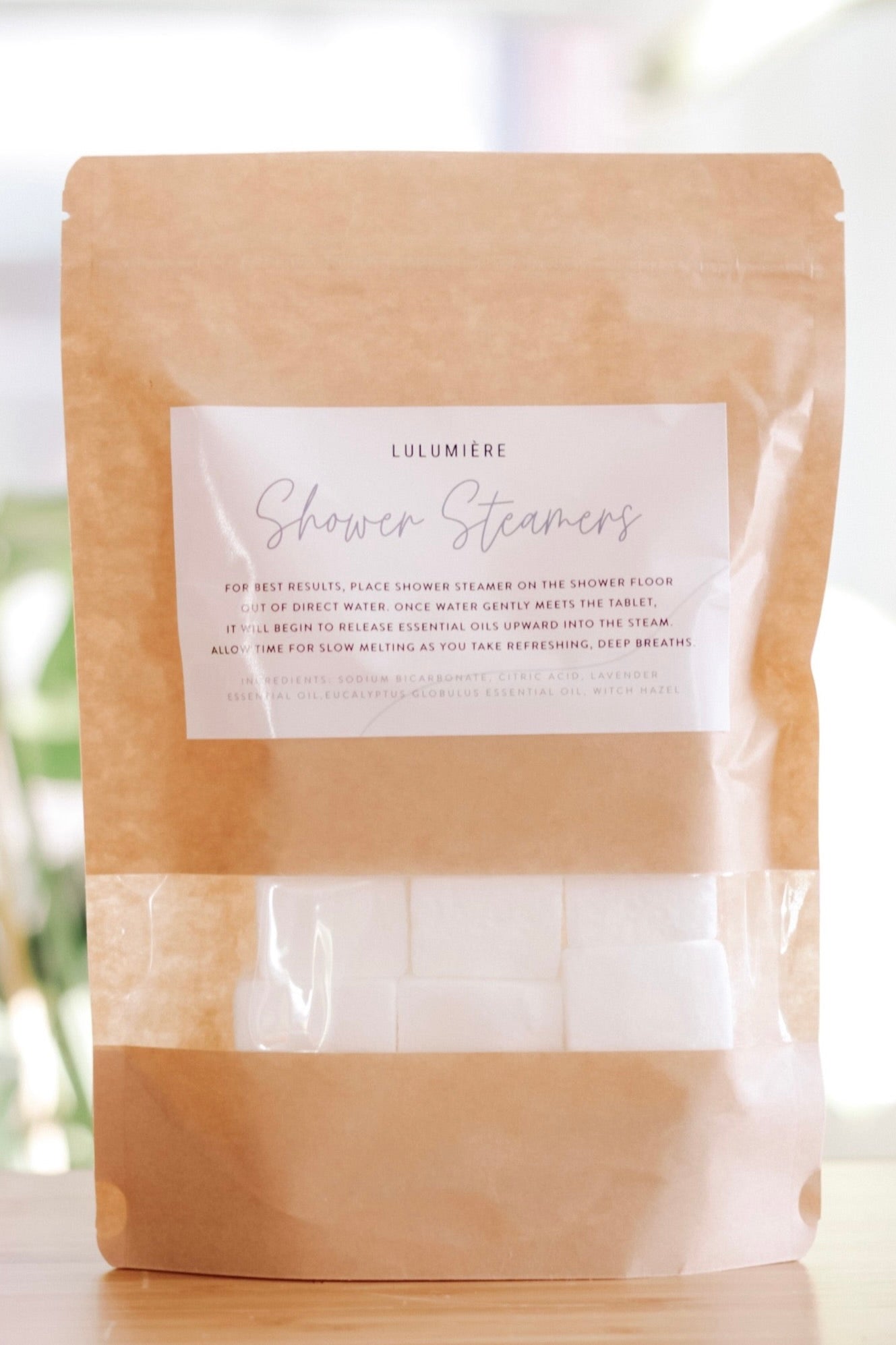 Brown paper pouches labeled 'Lulumiere Shower Steamers' with a clear window showing the white shower steamers inside.