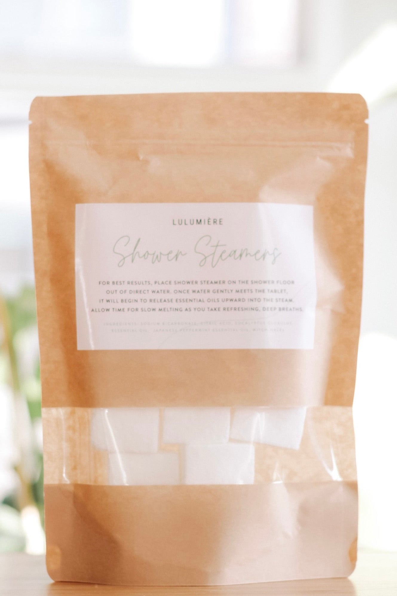 Two brown paper pouches labeled 'Lulumiere Shower Steamers' with a clear window showing the white shower steamers inside.