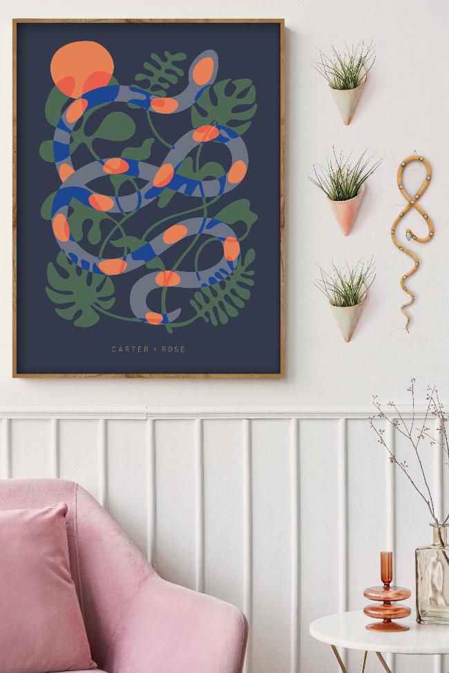 Styled framed dark blue poster with a modern snake and plant art design, and Carter + Rose text at the bottom. the snake is blue with green plants in the background and orange accents. Poster is styled with ceramic cones and snake in a sitting room setting. 