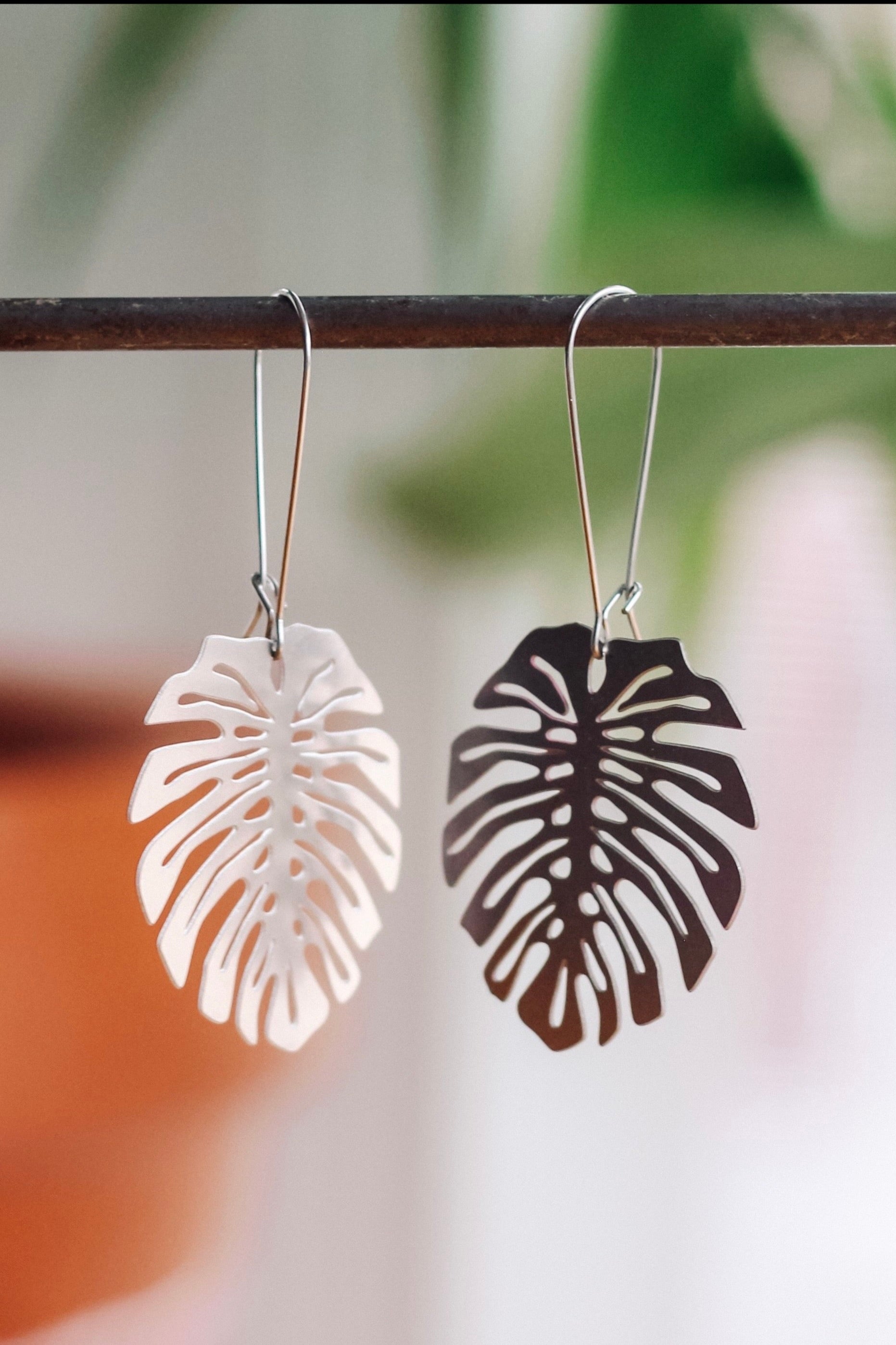 A pair of Monstera Deliciosa leaf-shaped earrings in stainless steel, displayed against a blurred background.
