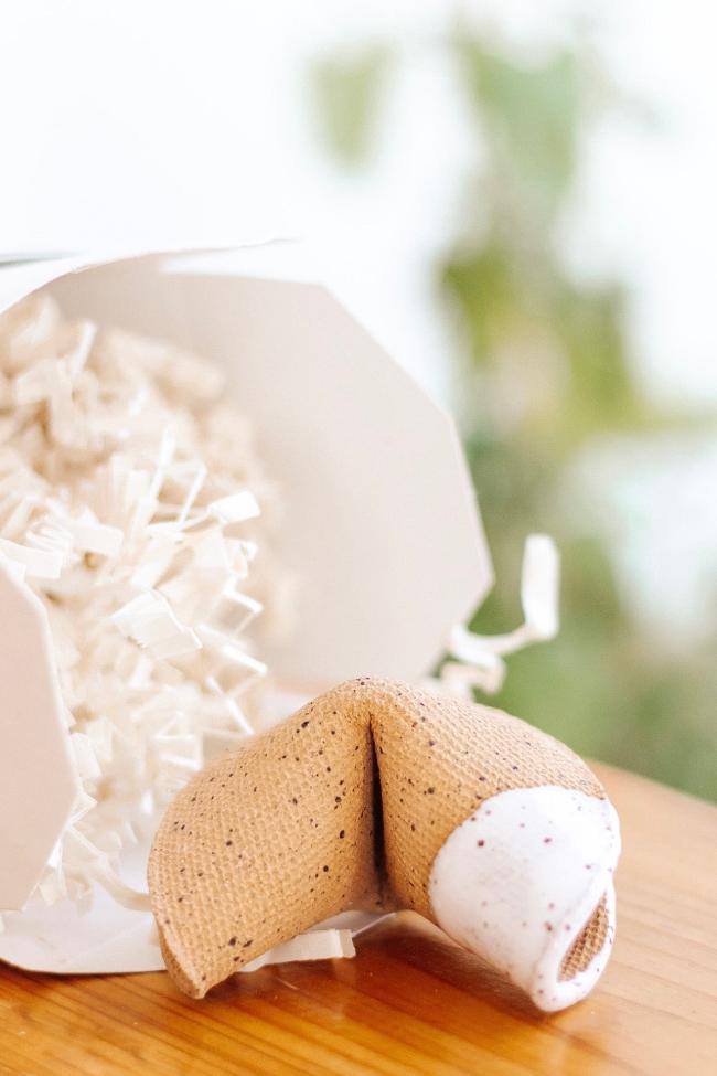 Styled ceramic speckled tan fortune cookie with one side dipped in an opaque white glaze next two a fortune cookie box