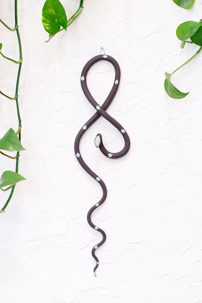Dark chocolate speckled clay vertical wall snake sculpture with light turquoise glazed head and dots. This colorway is Stevie. The top of the snake crosses like a figure eight and the tail hangs down in an organic s curve. It  hangs vertically from a small metal loop on a white wall with vining pothos. 