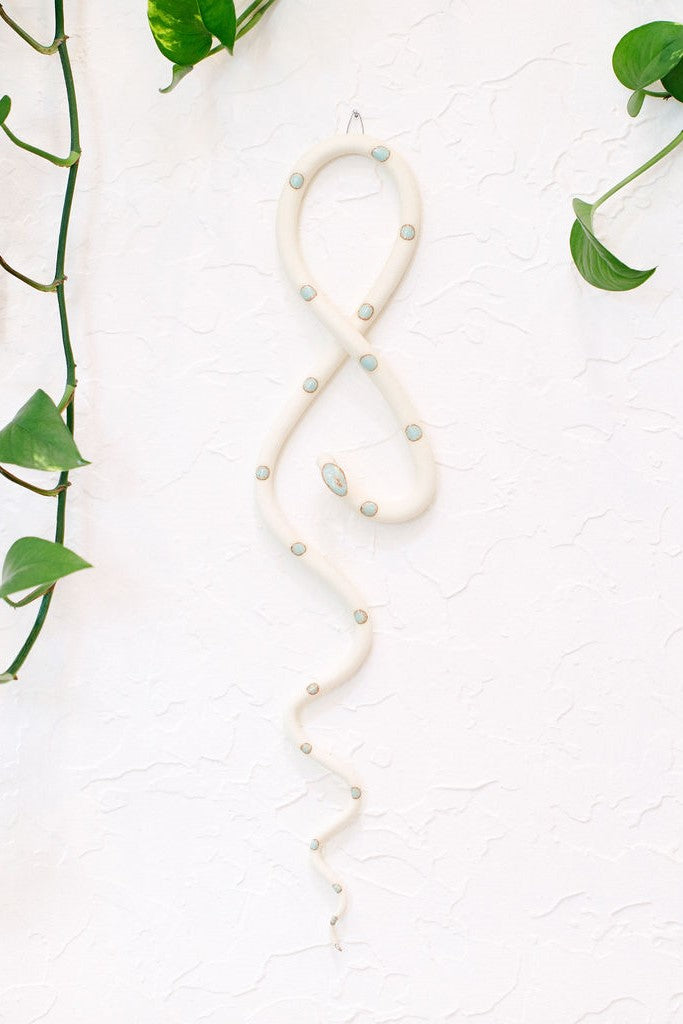 Ivory clay vertical wall snake sculpture with light turquoise glazed head and dots. This colorway is Sylvia. The top of the snake crosses like a figure eight and the tail hangs down in an organic s curve. It hangs vertically from a small metal loop on a white wall with vining pothos. 