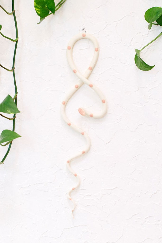 Ivory clay vertical wall snake sculpture with light pink glazed head and dots. This colorway is Sofie. The top of the snake crosses like a figure 8 and the tail hangs down in an organic s curve. It hangs vertically from a small metal loop on a white wall with vining pothos. 