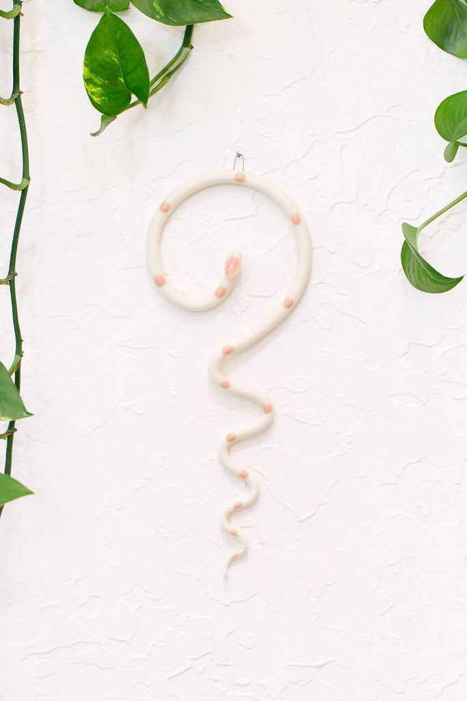 Simple contemporary ceramic snake wall decoration, with the top of the snake shaped loosely like a question mark and the tail hangs down in an organic s curve. This snake is Sofie, ivory with light pink glazed head and dots.
