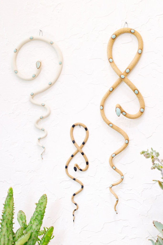 Desert curated set of three wall ceramic snakes. From left to right hanging on a wall a medium Sylvia wall snake (white clay, turquoise dots), a Sammy mini wall snake (tan clay, black dots) and a Selma wall snake (tan clay, turquoise dots). 