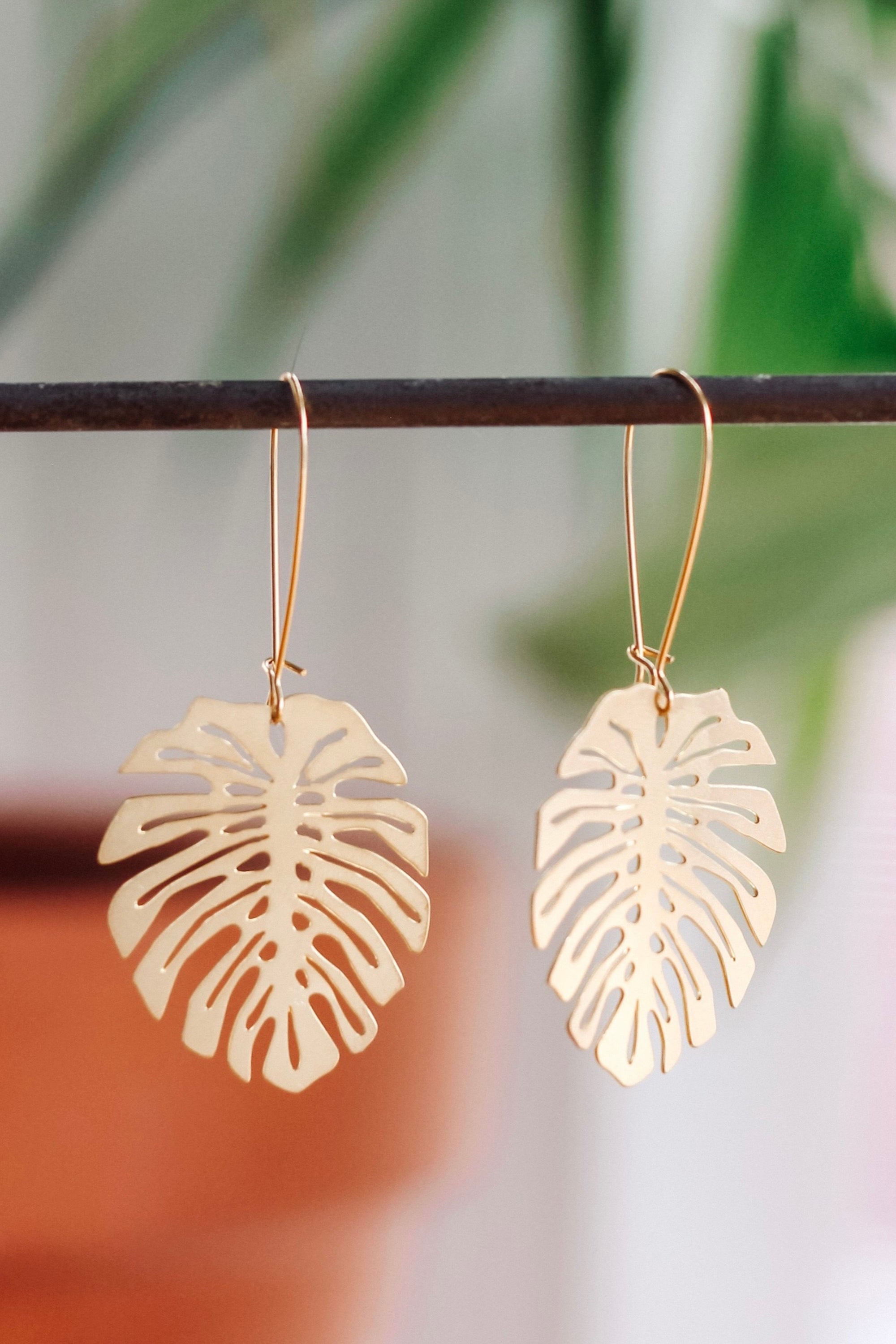 A pair of brass Monstera Deliciosa leaf-shaped earrings, displayed against a blurred background.