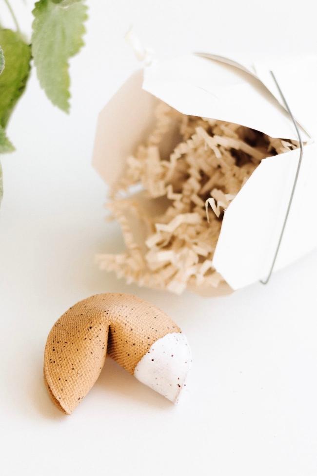 A handmade ceramic fortune cookie by Carter + Rose, displayed next to a fortune cookie box for packaging on a white background with some green leaves at the top left corner.