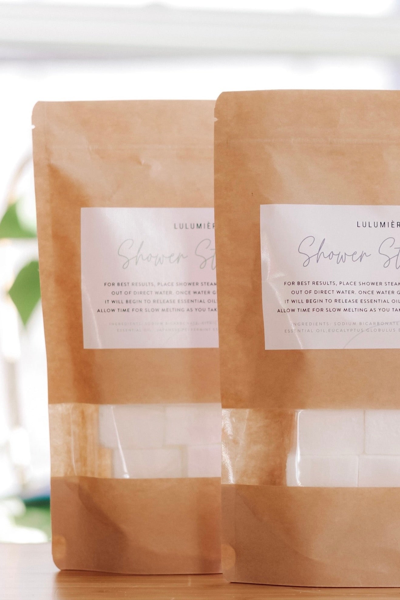 Two brown paper pouches labeled 'Lulumiere Shower Steamers' with a clear window showing the white shower steamers inside.