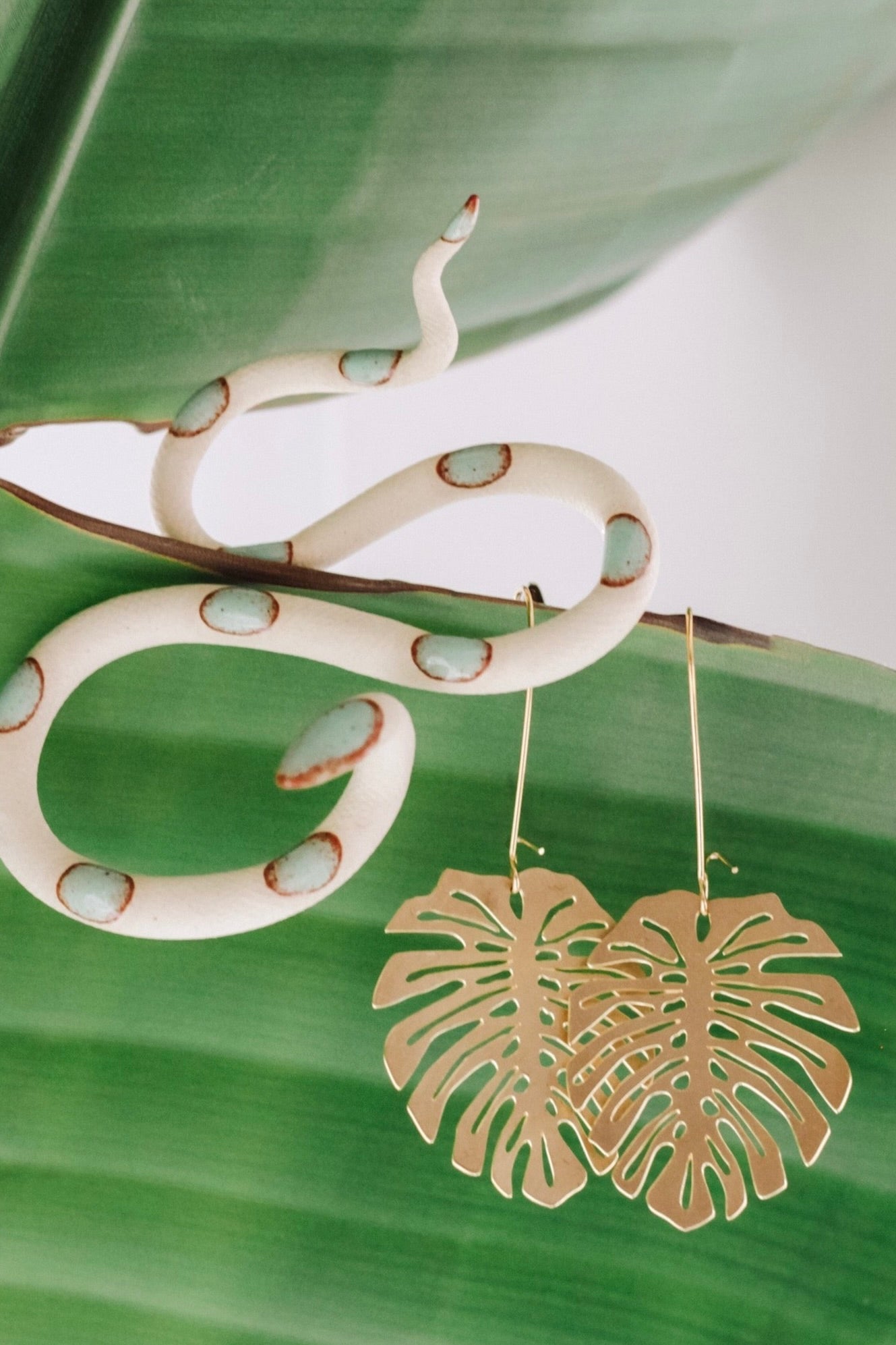 White ivory clay snake sculpture with green dots sitting next to gold monstera shaped earrings