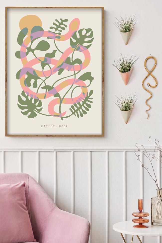Styled framed ivory poster with a modern snake and plant art design, and Carter + Rose text at the bottom. The snake is pink, with green plants in the background and yellow accents. Poster is styled with ceramic cones and snake in a sitting room setting. 