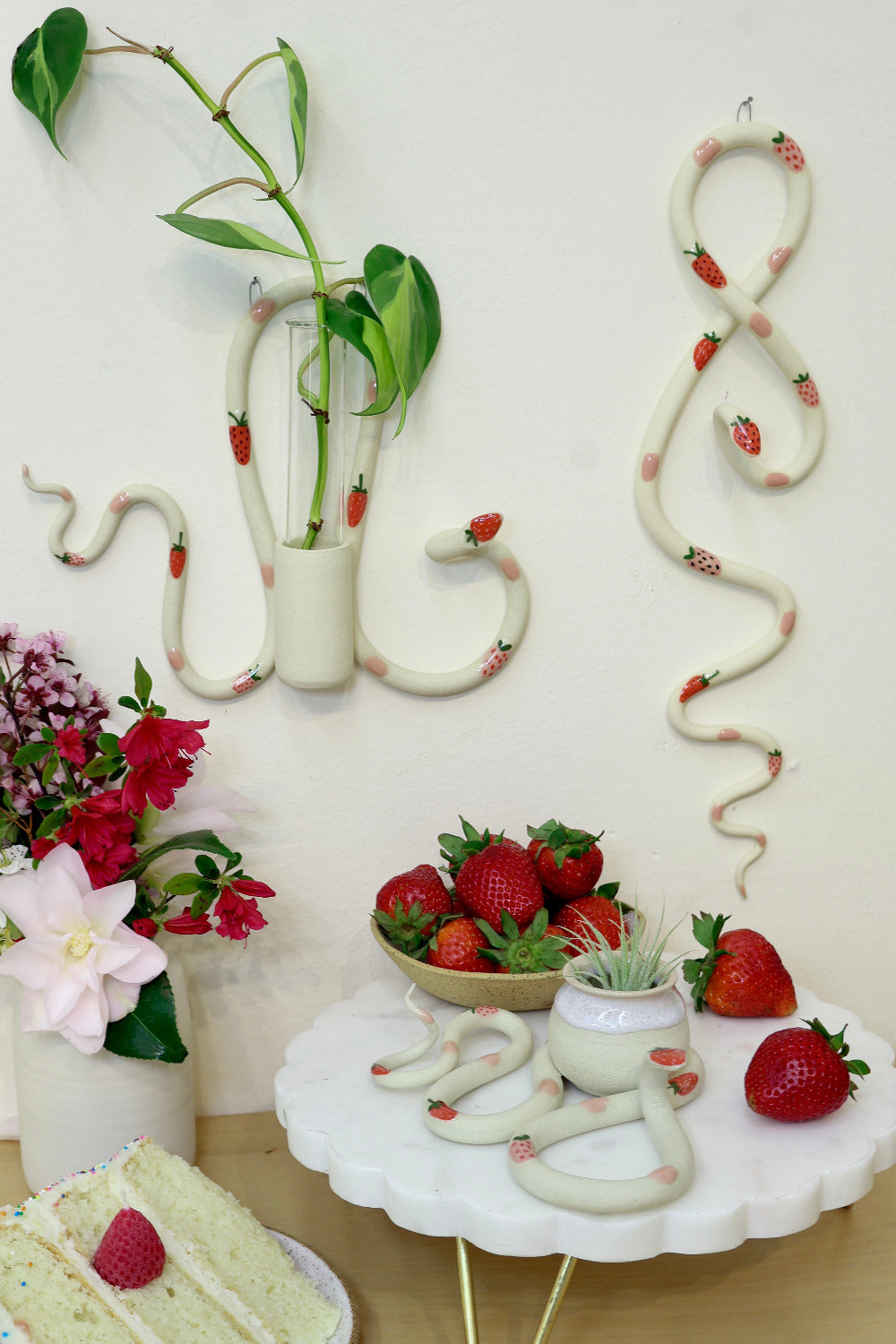 A whimsical display featuring ceramic snake-shaped wall vases and decorative pieces, adorned with red strawberry accents and pink polka dots. Fresh strawberries in a bowl and a slice of cake with a strawberry on top complete the charming arrangement.