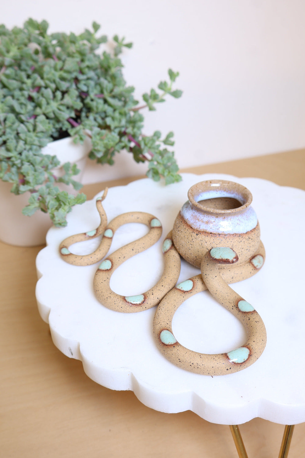 A speckled tan ceramic snake-shaped sculpture with with green dots and a small bud vase on top. The vase is also speckled tan with a transparent white glazed top. This colorway is Selma. It's styled next to a houseplant.