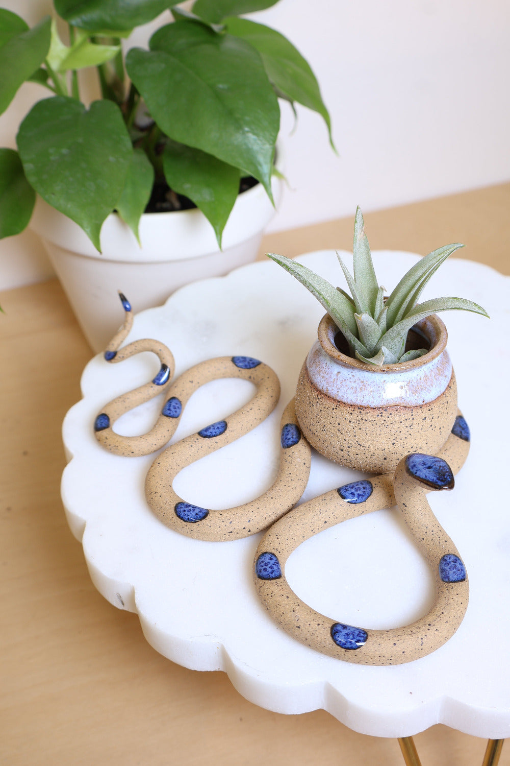 A speckled tan ceramic snake-shaped sculpture with with blue dots and a small bud vase attached in one of the snakes curves. The vase is speckled tan clay with a opaque white glazed top. This colorway is Selena. It's styled with a houseplant and an air plant in the vase.
