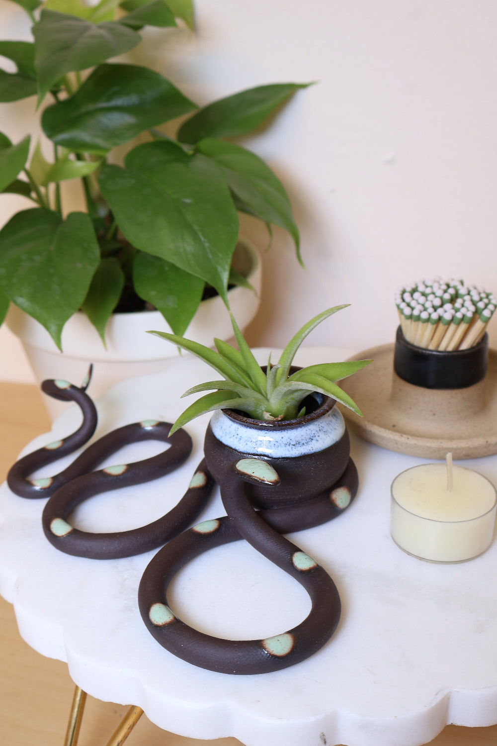A black ceramic snake-shaped sculpture with with green dots and a small bud vase on top. The vase is also black with a transparent light blue glazed top. This colorway is Stevie. It's styled next to a houseplant and matches.