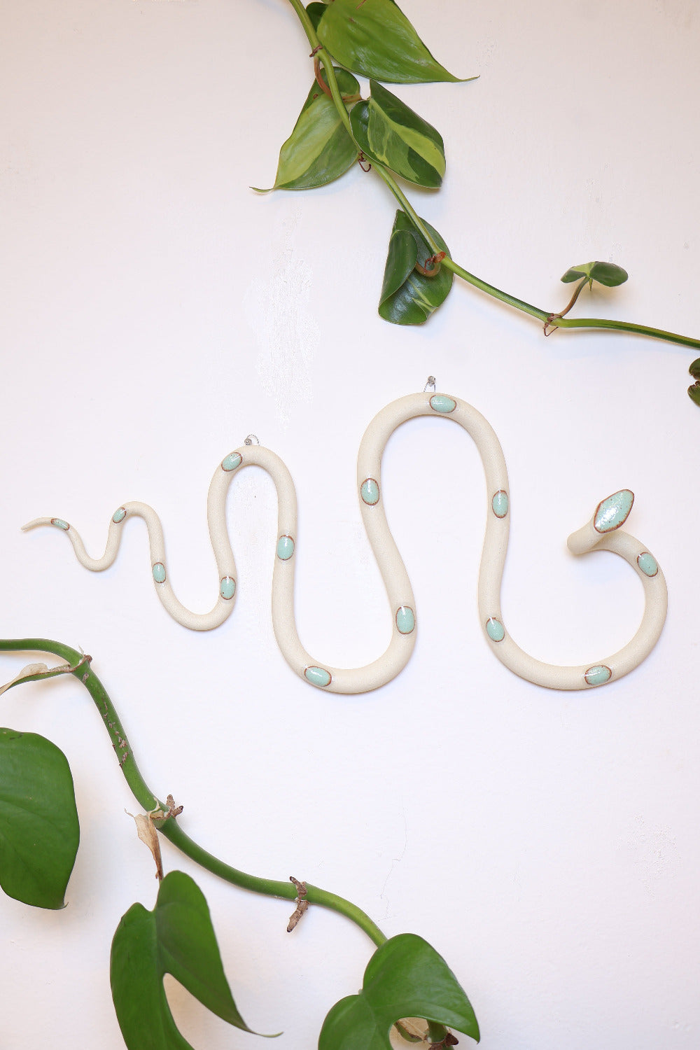 Horizontal wavy ceramic wall snake in the Sylvia colorway, ivory clay base and light turquoise dots. It's hanging on a white wall with two small nails by its two small metal hooks.