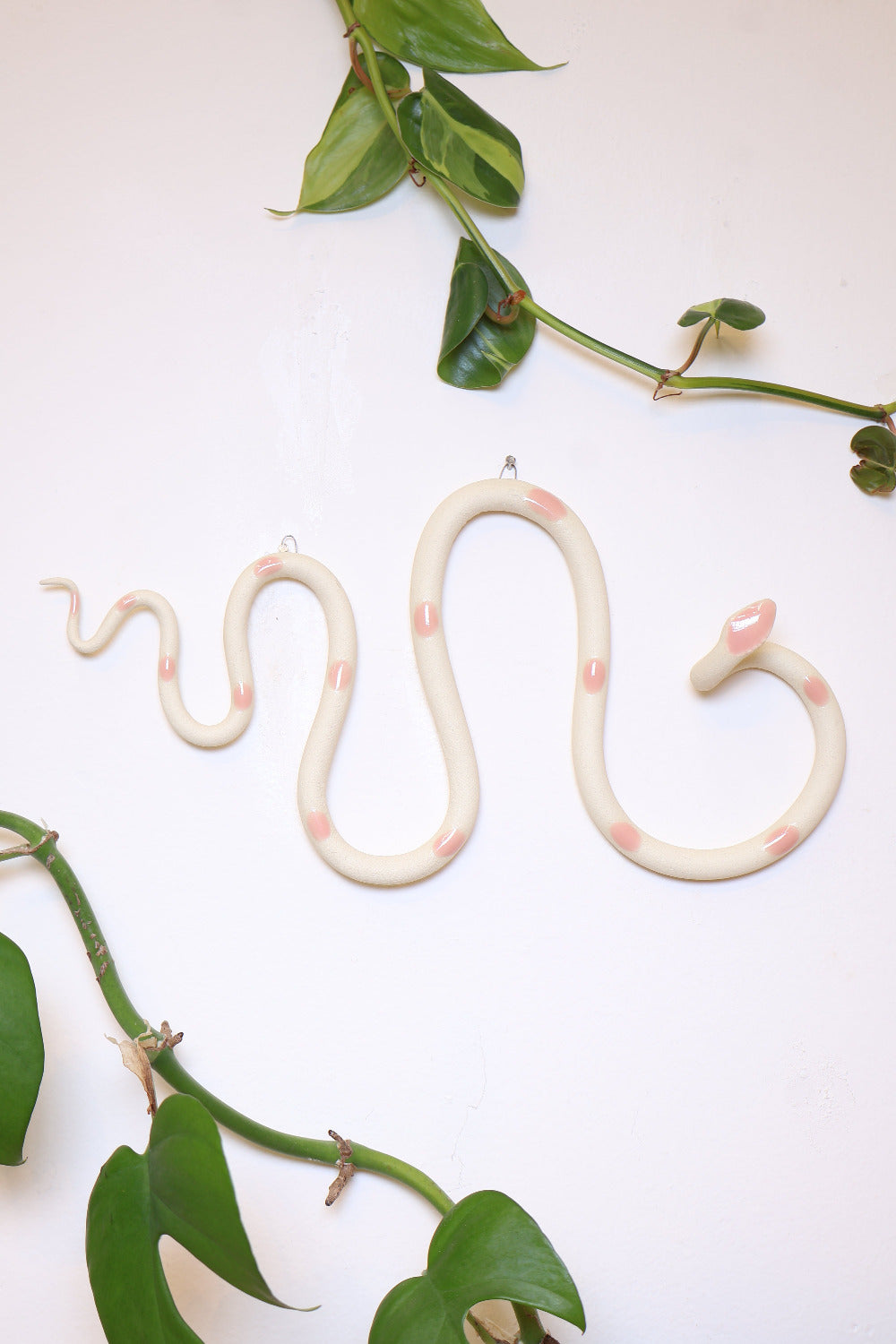 Horizontal wavy ceramic wall snake in the Sofie colorway, ivory clay base and light pink dots. It's hanging on a white wall with two small nails by its two small metal hooks.