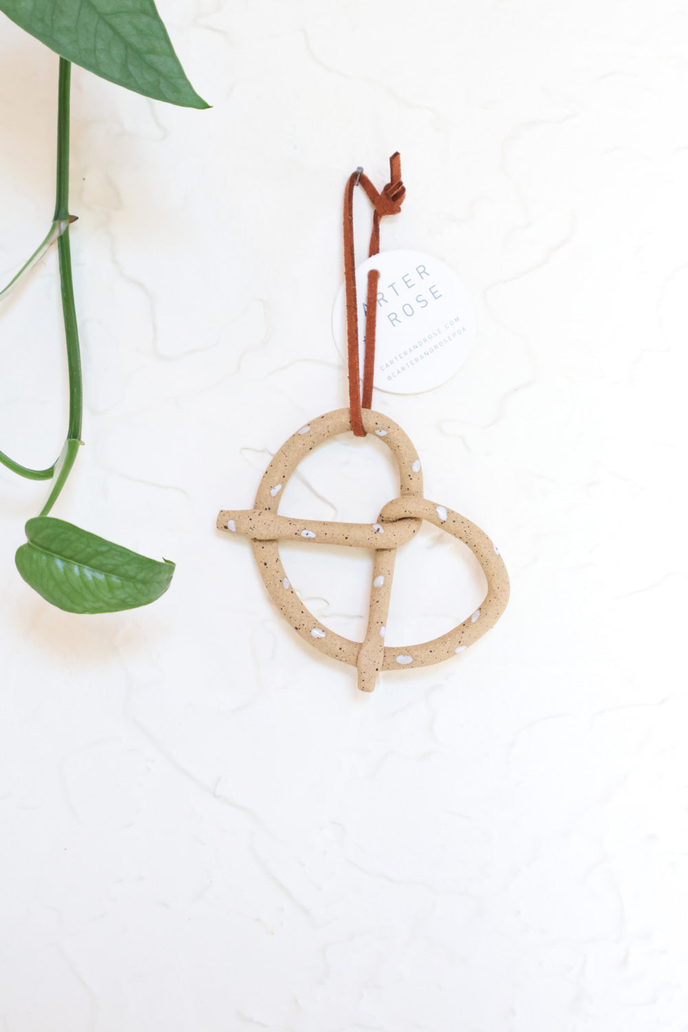 A tan ceramic pretzel-shaped ornament hanging against a wall, with a green leaf partially visible at the top.