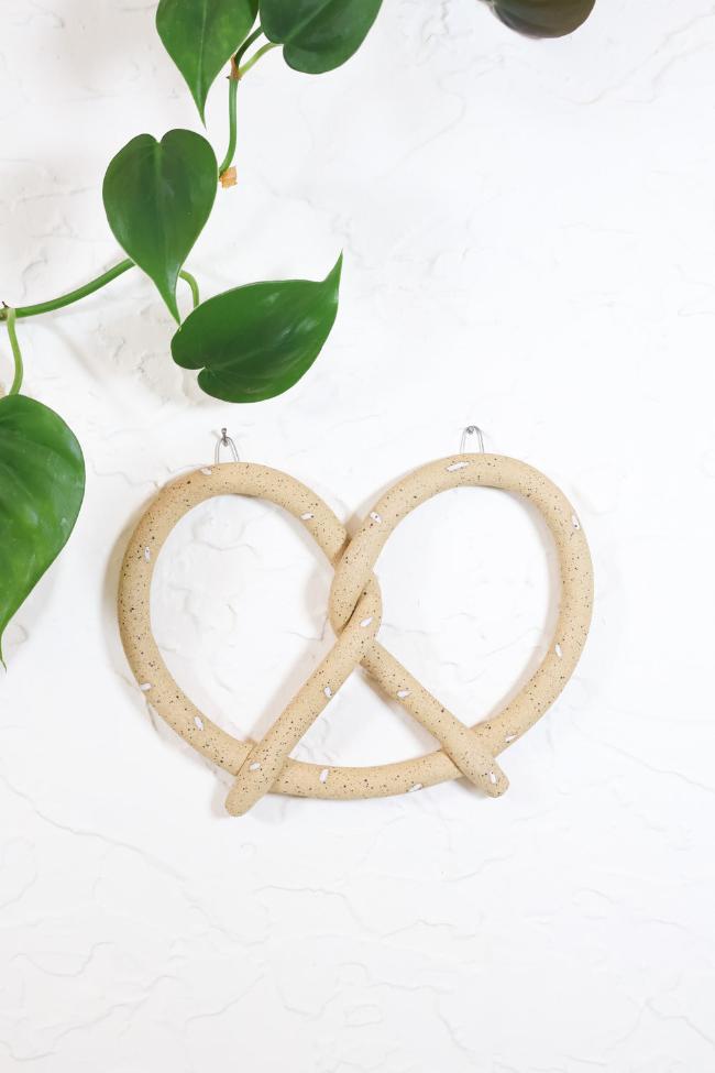 A tan ceramic pretzel wall hanging with white speckled salt sprinkles. The pretzel is hanging from two small silver hooks on a white wall with a vining green plant.