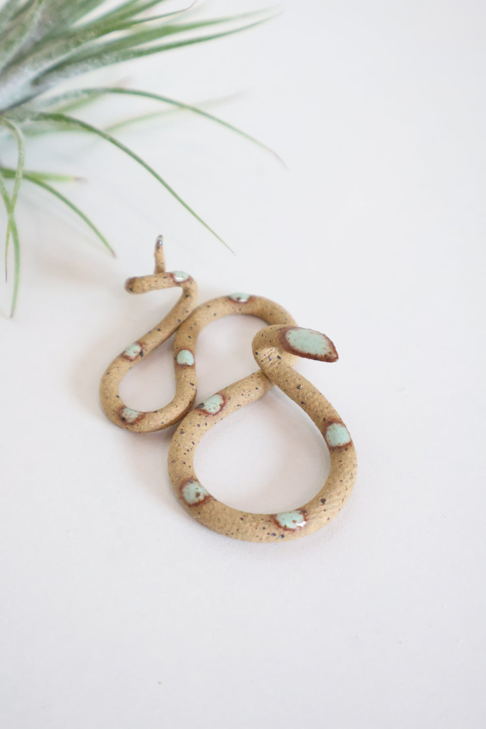 Itty bitty organic s-shaped pottery snake sculpture in the colorway Selma, speckled tan clay body with a light turquoise glazed head and evenly spaced glazed dots.