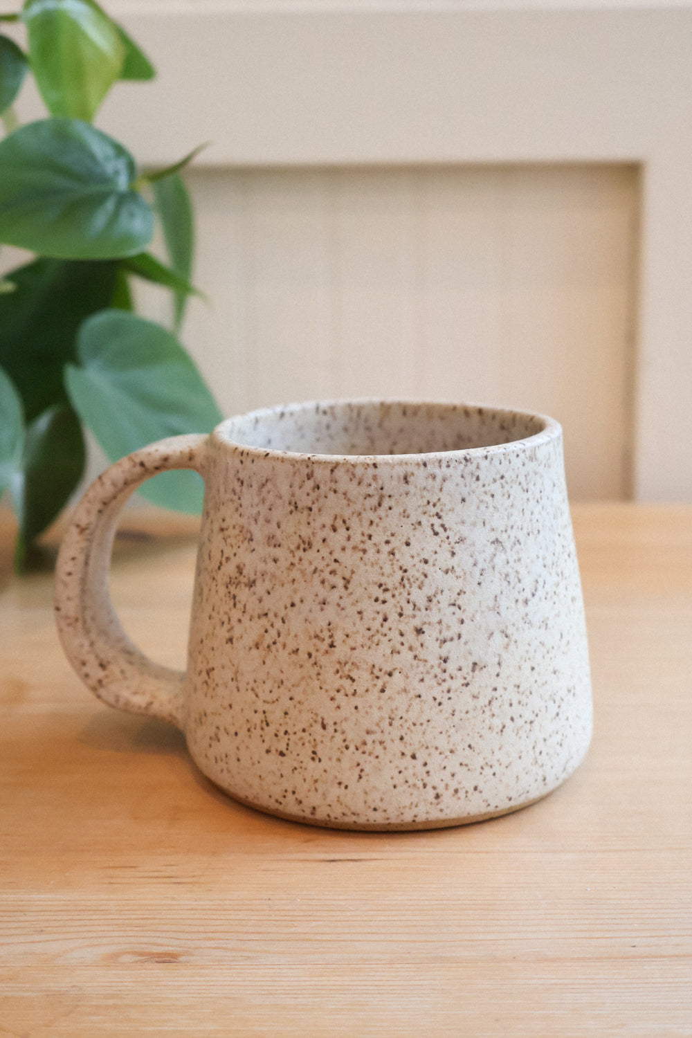 Speckled white and tan ceramic mug on a wooden surface with a plant in the background