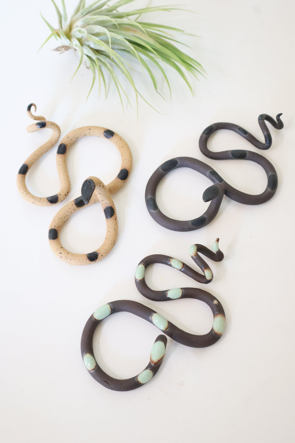 Forest curated set of three small ceramic snakes in the following colorways, Saul (black clay with black glaze dots), Stevie (black clay with light turquoise glaze dots), and Sammy (speckled tan with black glaze dots).