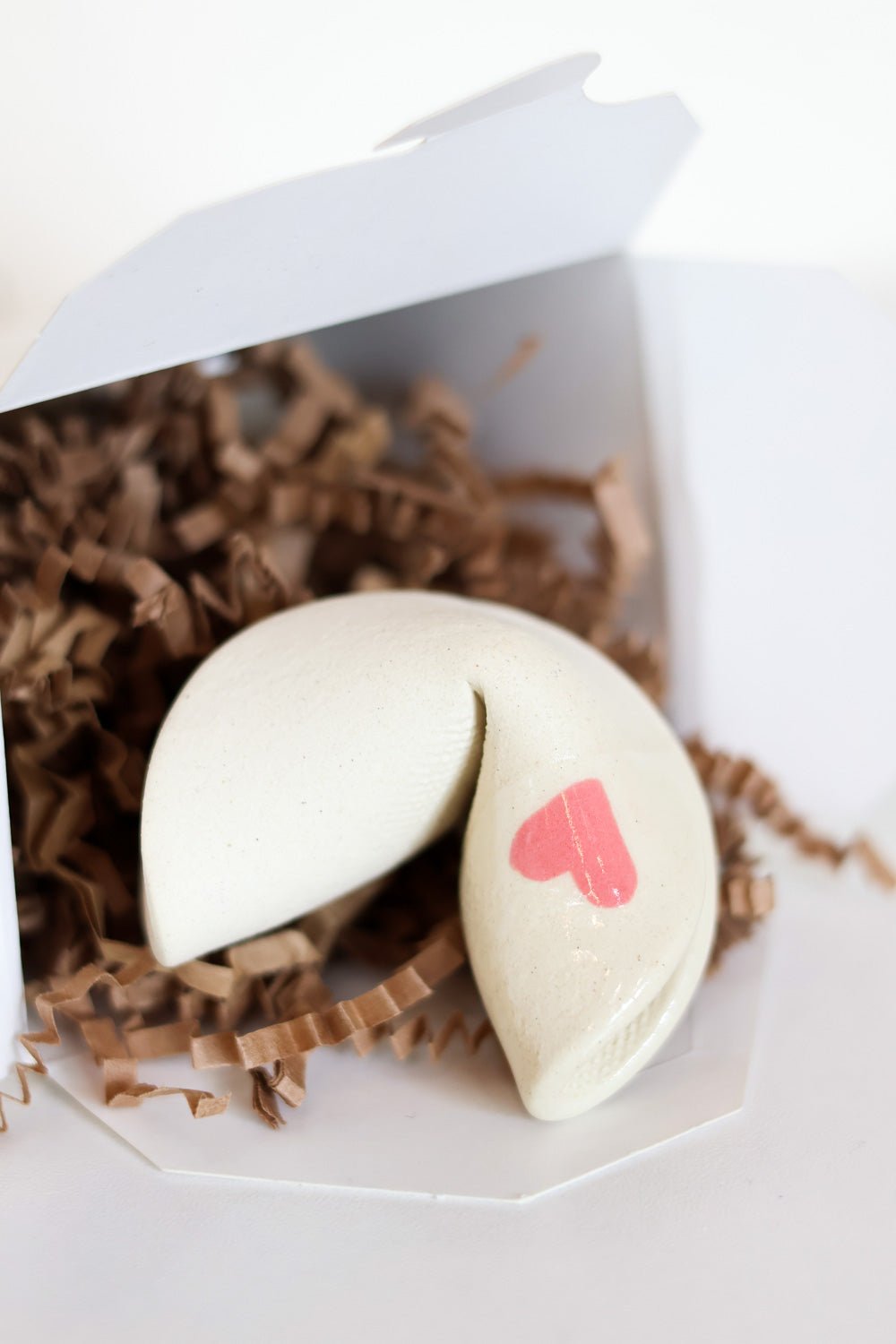 Ceramic fortune cookie made from ivory clay with a little pink heart on one side inside a fortune cookie box