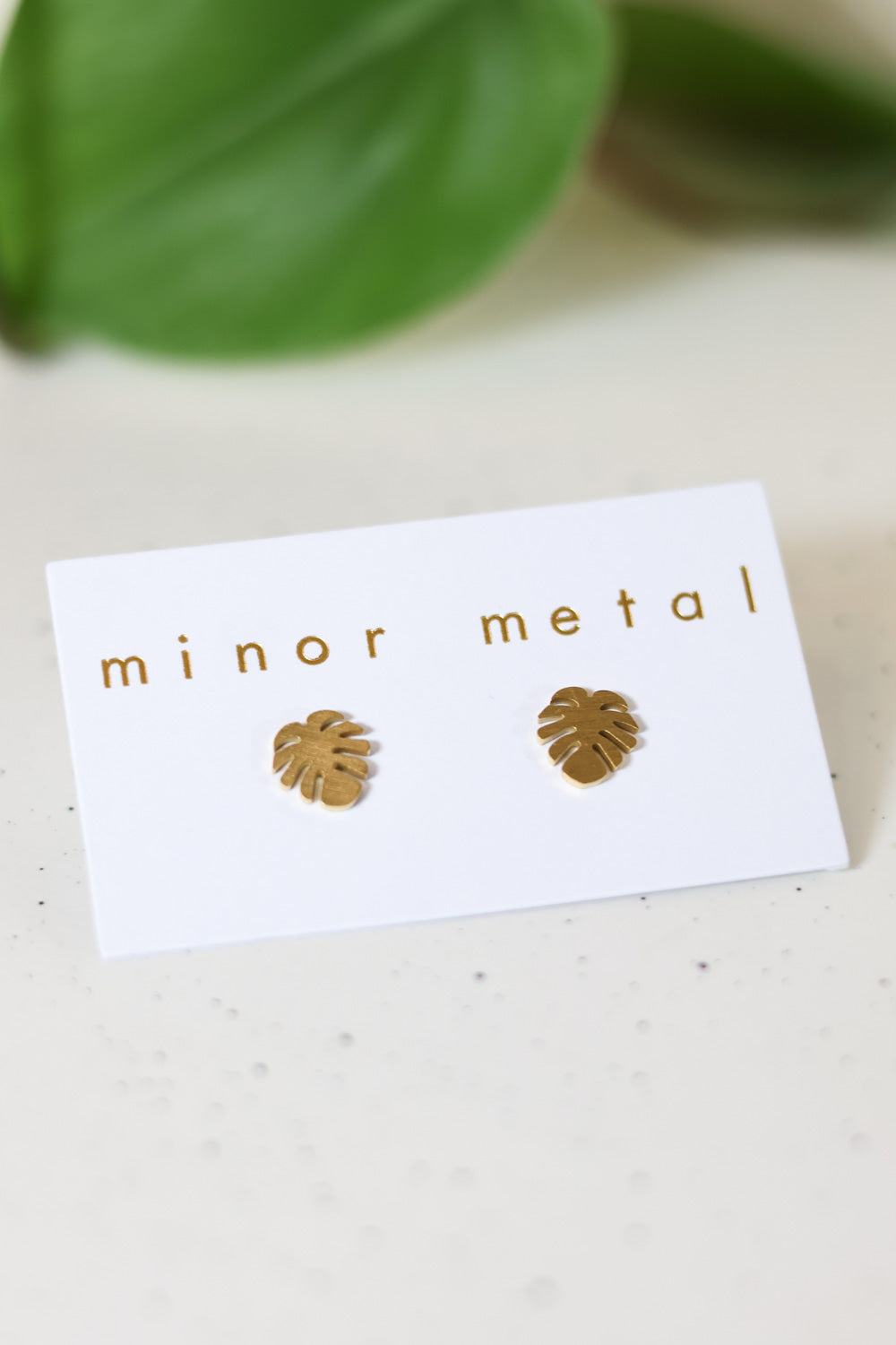 A pair of small monstera leaf-shaped gold stud earrings on a white card with 'minor metal' written in gold lettering.