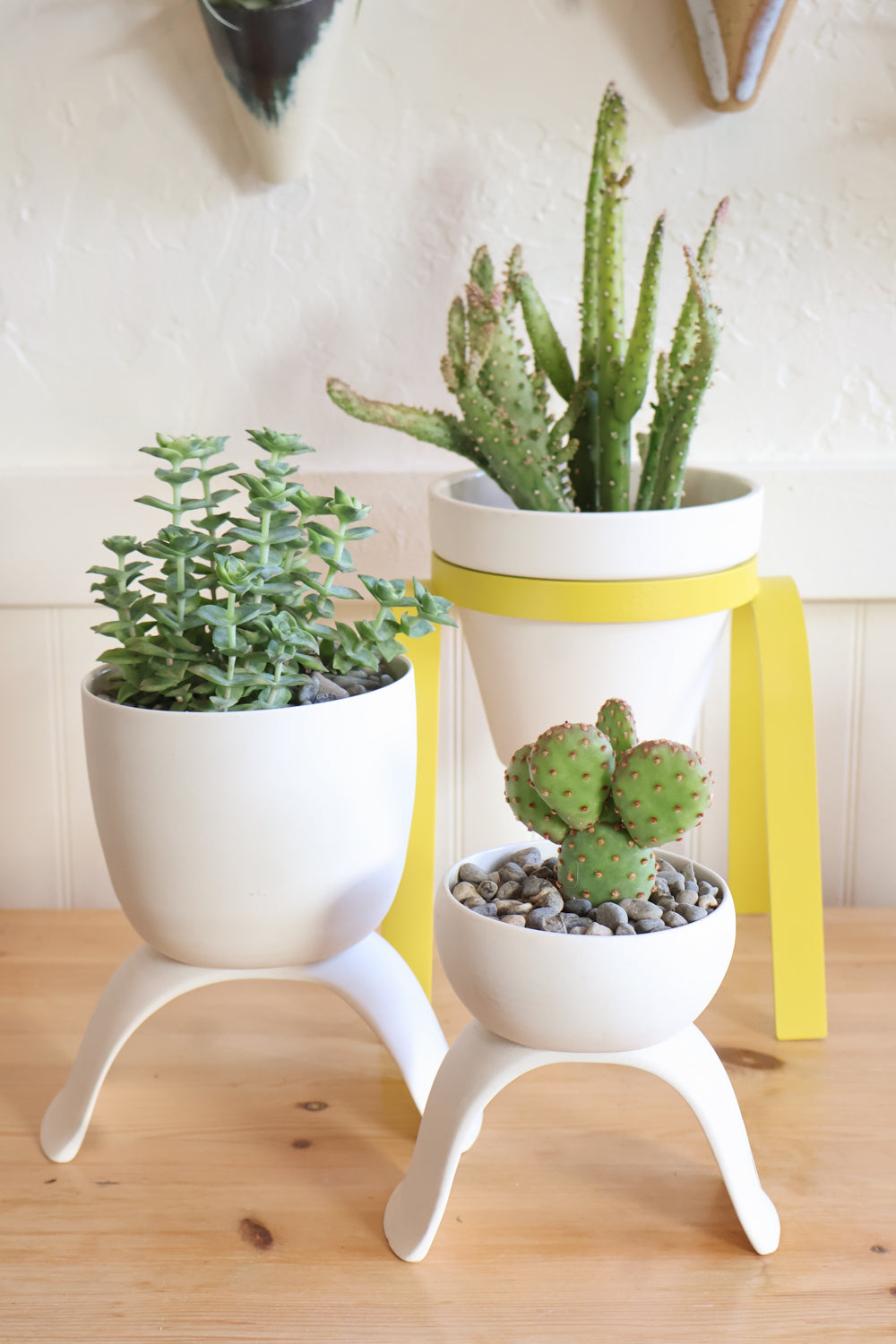 Two white porcelain high-rise plant pots in different sizes and a yellow metal raised plant stand with a white pot all with potted succulents. The pots are displayed on a wooden surface with a decorative ceramic cones hanging in the background.