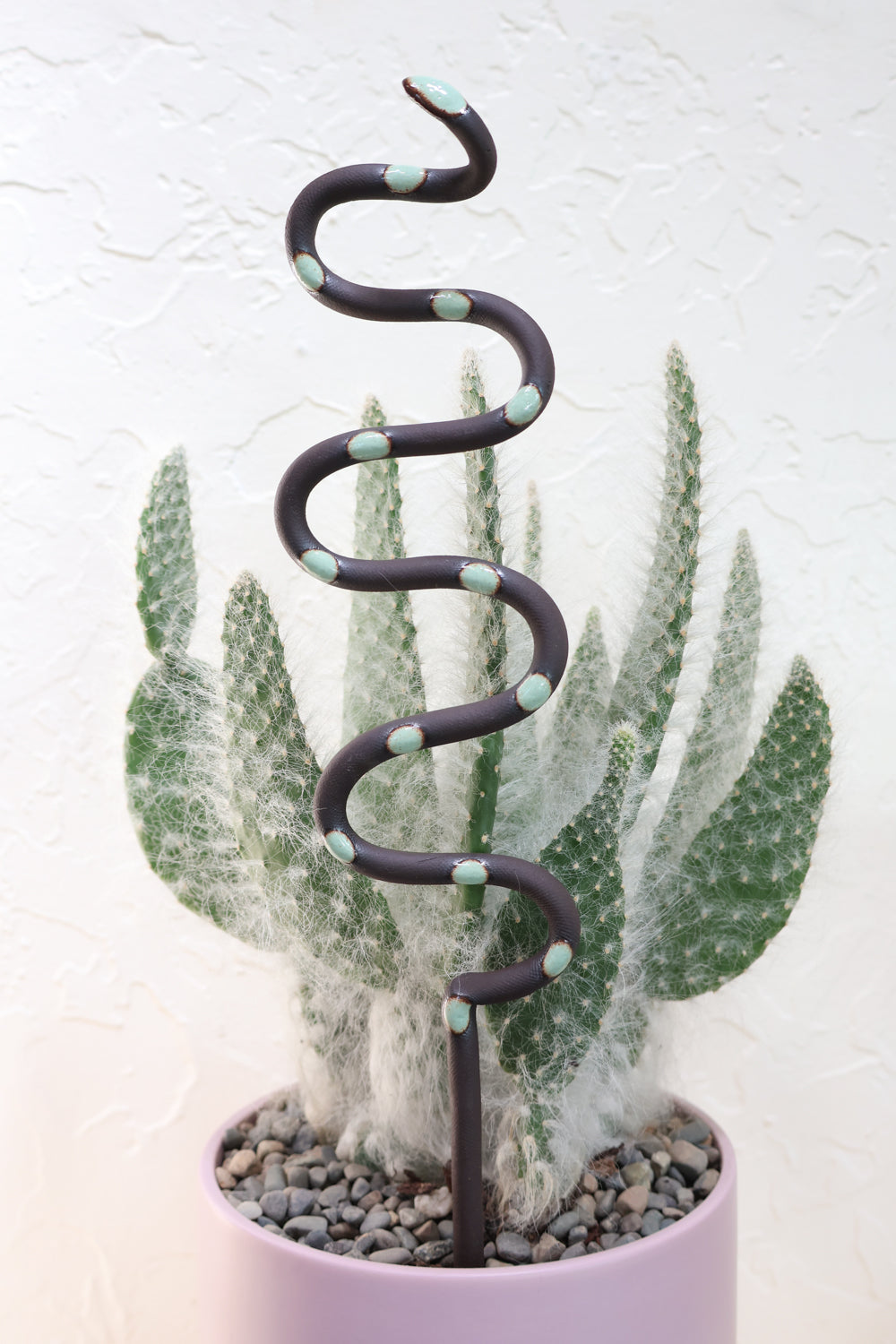Stevie colorway clay plant stake in a pot with a cactus. This snake is black with green spots. 