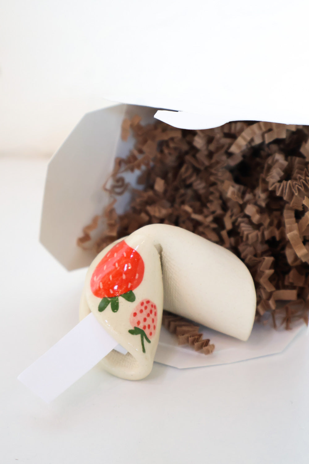 Ceramic fortune cookie made from ivory clay with a red and pink strawberry decal on one corner in a fortune cookie box.