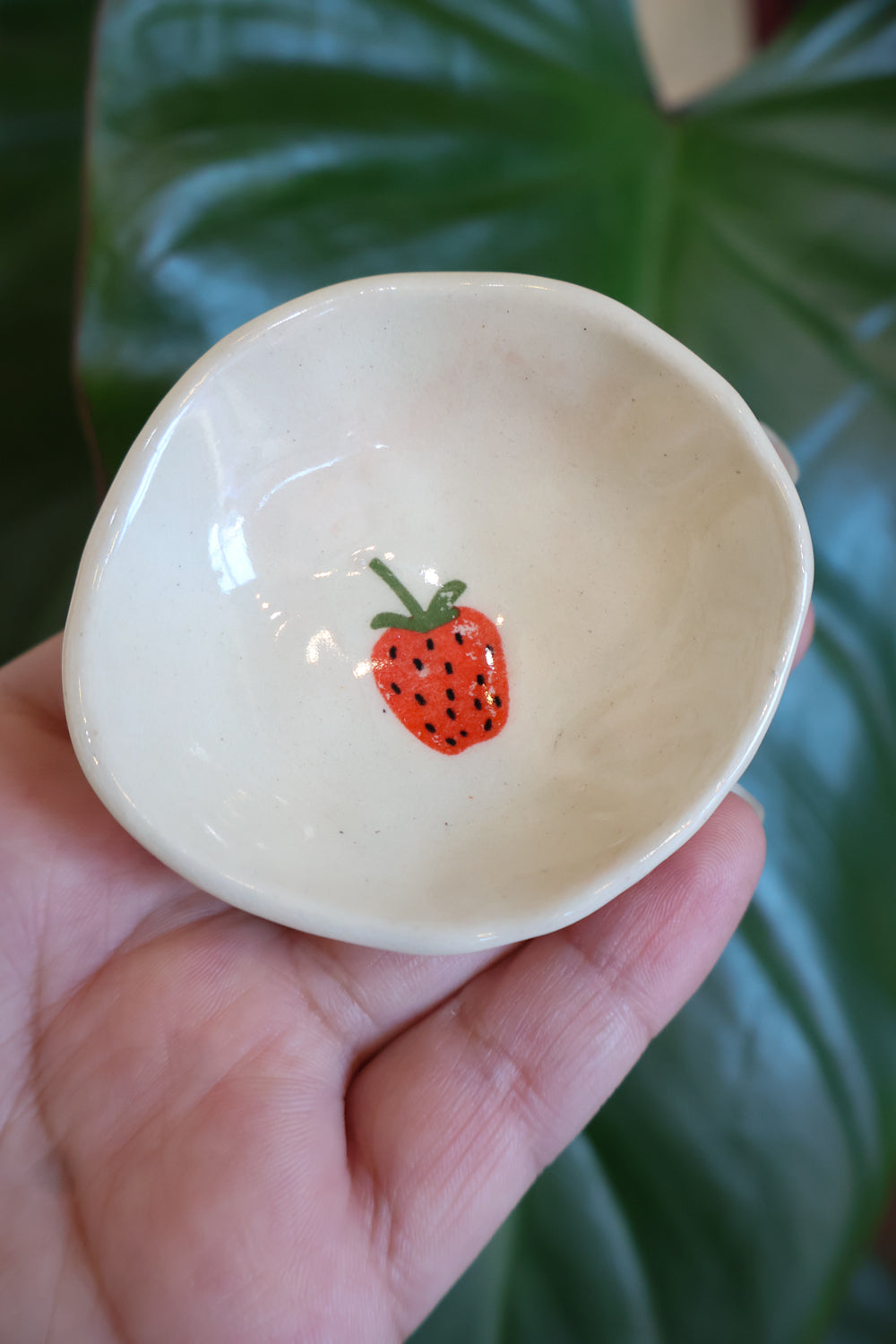 Small white bowl with a single strawberry held in a hand against a green leafy background