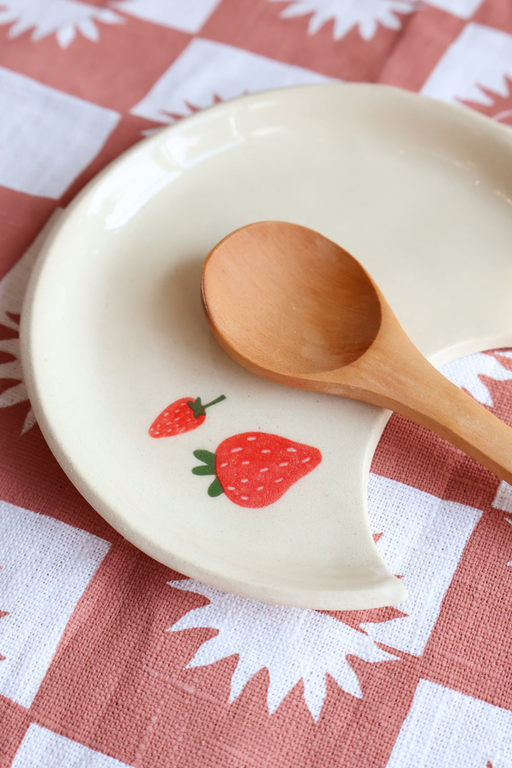 White ceramic spoon rest with two strawberry decals displayed with a wooden spoon on a red and white checkered tablecloth.