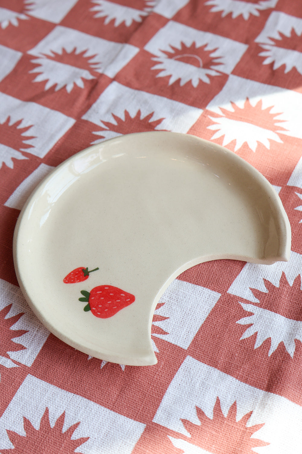 White ceramic spoon rest plate with strawberry design on top of a red and white checkered tablecloth