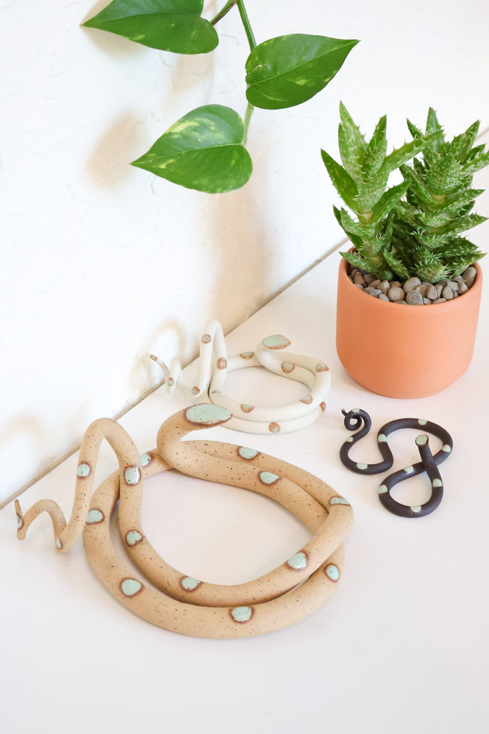 Three different sizes of clay snakes. One large round speckled tan snake with green spots in the foreground. A small black clay snake with green dots to the right of it, and a smaller round white snake with green dots to the left. In the background is a little potted succulent. 