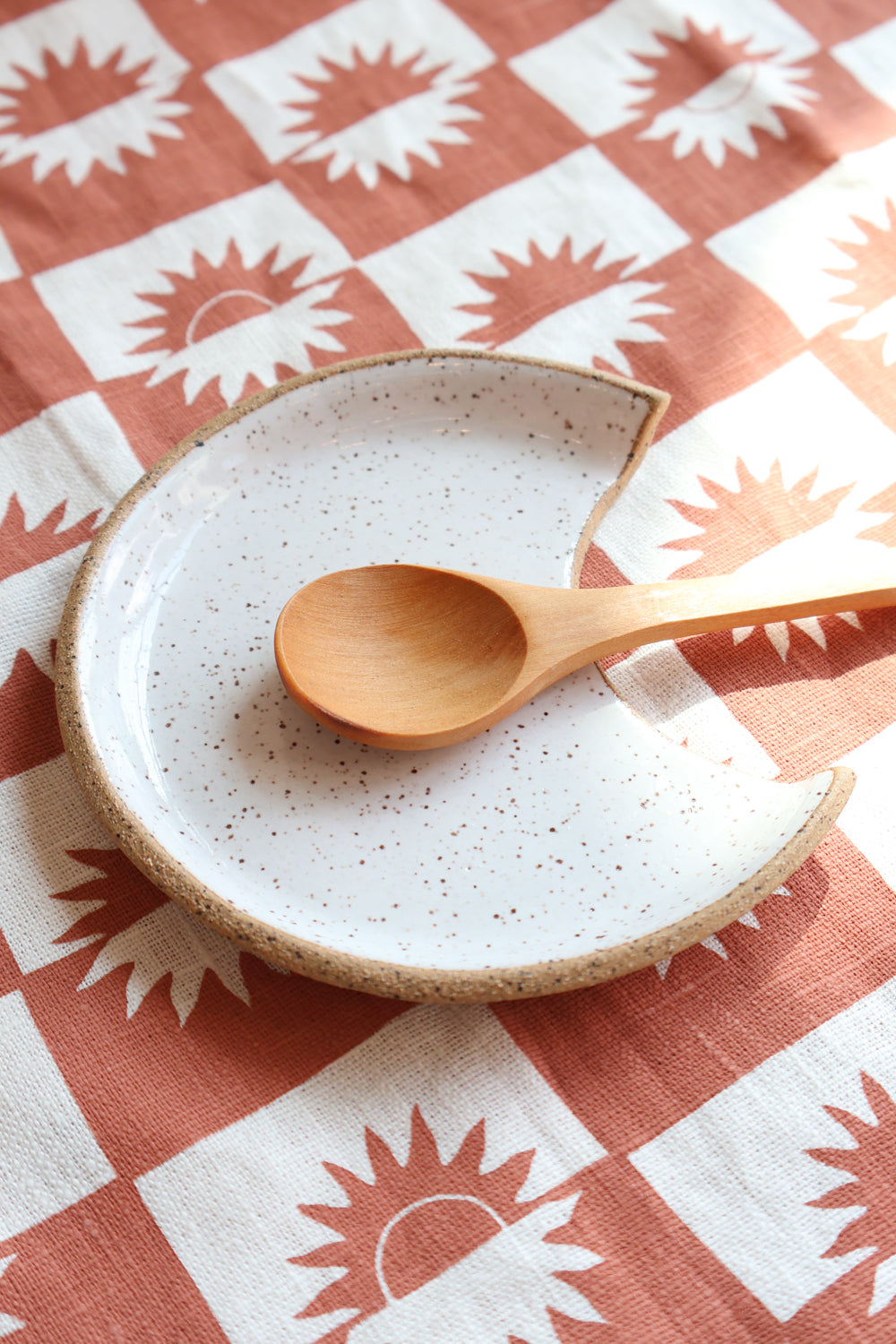 White speckled ceramic spoon rest displayed with a wooden spoon on a patterned fabric background