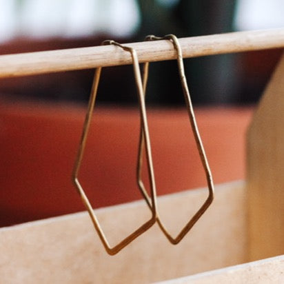 A pair of brass geometric hoop earrings hanging from a wooden earring holder, with a plant in the background.